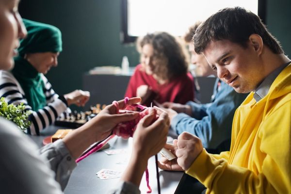 A group of people at a long table doing crafts and playing games. In focus a young man with short, dark hair wearing a yellow sweater looks at the knitting another person at the table is working on.